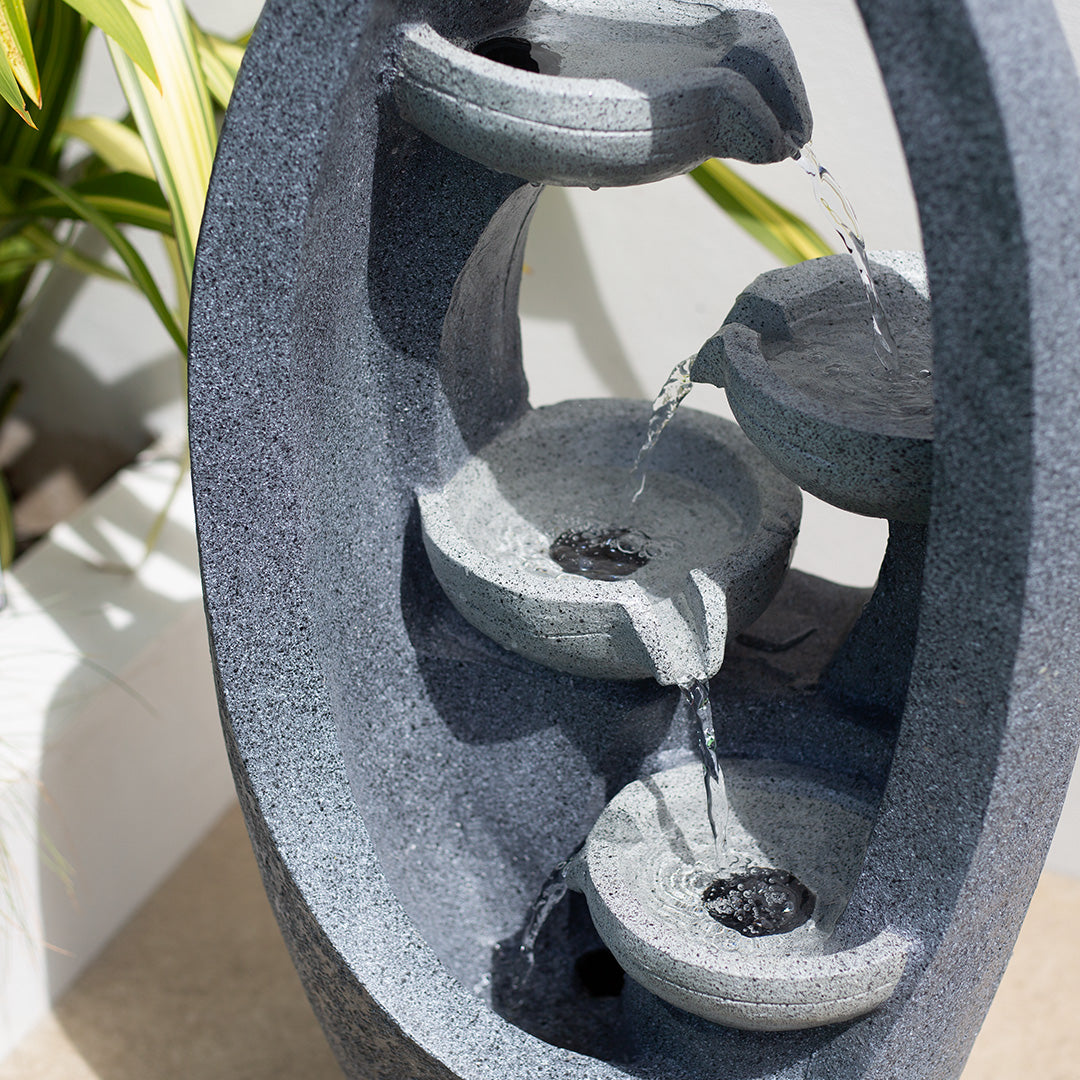 The La Hacienda Sparkling Pours incl LEDs is a tiered stone solar patio fountain with an oval frame, designed with four small cascading bowls. Water gently flows from the top to the bottom bowl using a recirculating pump, and lush green leaves can be partially seen in the background.