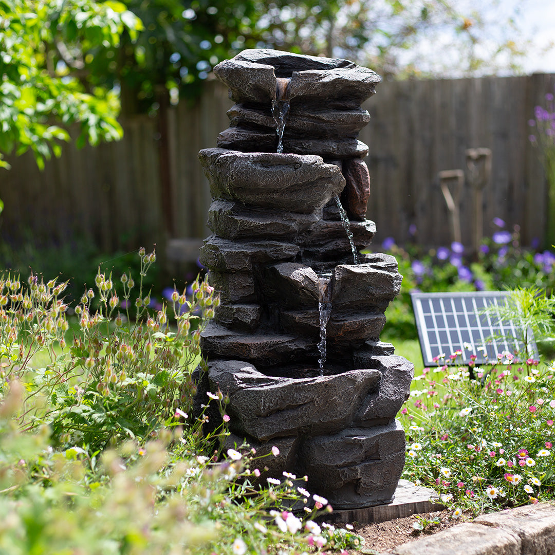 A Hamsterley Springs water feature by La Hacienda, with LED lights, mimics a rocky waterfall as it cascades down multiple levels in the garden. Powered by a solar-powered recirculating pump, it is surrounded by green plants and small flowers. Nearby, a solar panel can be seen alongside a wooden fence and garden tools.