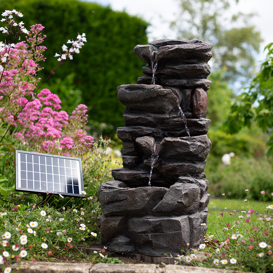 The garden is adorned with the Hamsterley Springs incl LEDs, a magnificent solar-powered cascading water feature from La Hacienda. This multi-tiered fountain, equipped with a recirculating pump, allows water to flow gracefully. It is beautifully complemented by pink and white flowers and lush greenery, while a nearby solar panel provides the energy for its captivating allure.