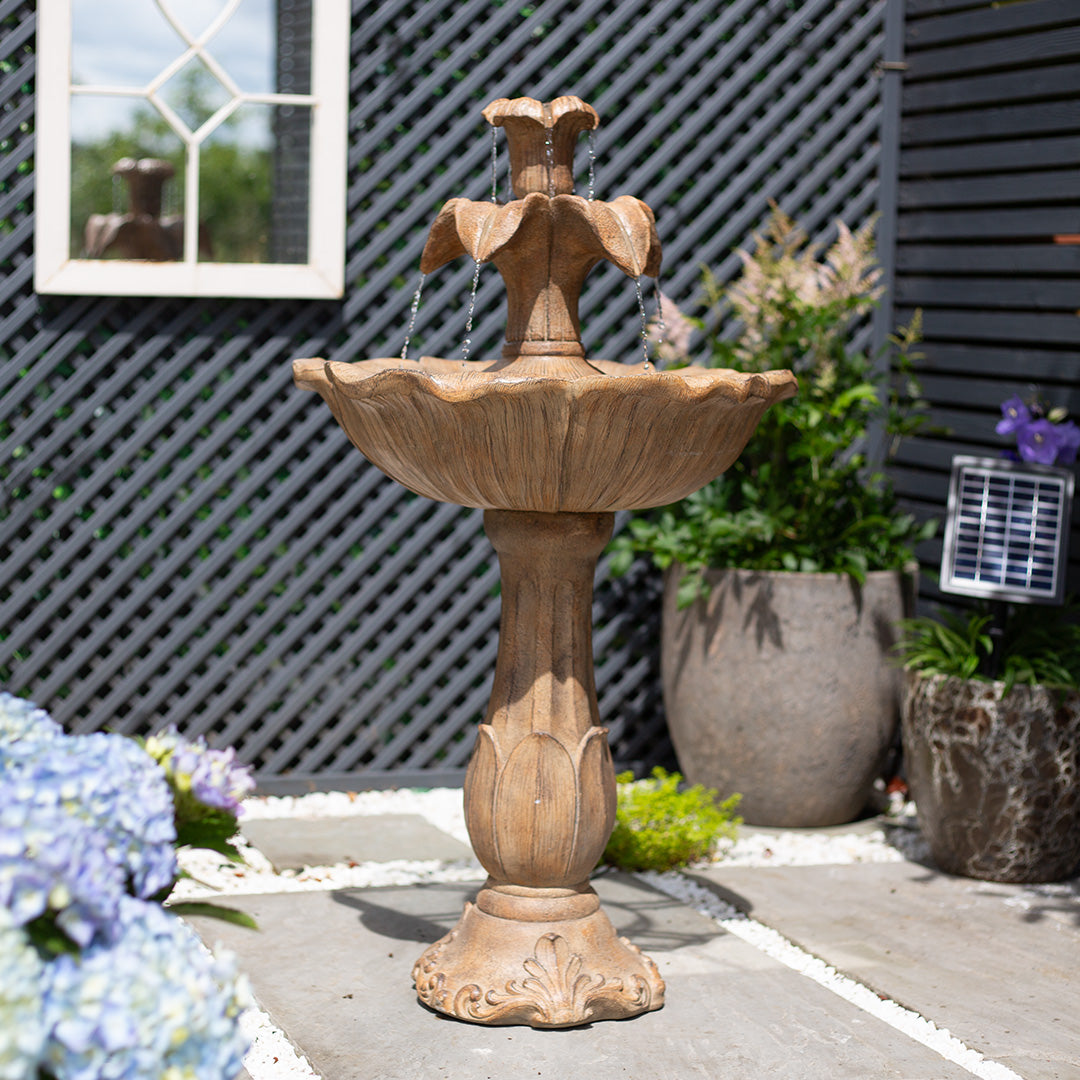 The tiled patio becomes a captivating outdoor retreat as the La Hacienda Emily Tiered Fountain, a garden centerpiece, stands amidst potted plants. This artistic stone fountain features solar-powered technology that elegantly cascades water from its two tiers. A lattice fence and window capture the peaceful ambiance of the scene.