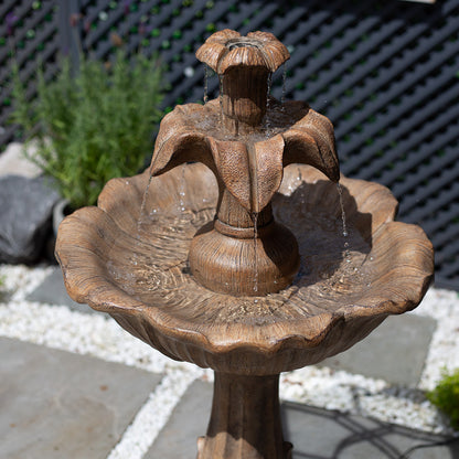 The Emily Tiered Fountain by La Hacienda is a rustic brown stone solar-powered water feature with water cascading from a flower-like top into a scalloped basin, perfectly set on a patio. The scene is enhanced by lush greenery and a lattice fence in the background, making it an ideal garden focal point.