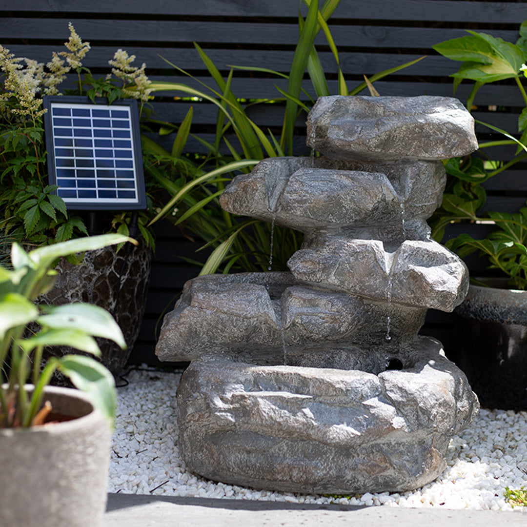 The garden features the La Hacienda's Cascading Rocks incl LEDs, a solar-powered fountain with a natural stone finish. Water gently cascades over its stacked formations, surrounded by lush green plants and white pebbles that enhance the serene and enchanting atmosphere.