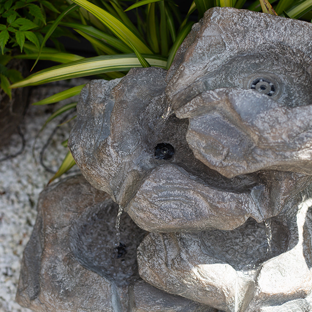 The "Cascading Rocks incl LEDs" by La Hacienda is a decorative solar-powered garden fountain featuring water cascading down three tiers, surrounded by green plants. It boasts a natural stone appearance with a rocky texture subtly illuminated by sunlight.