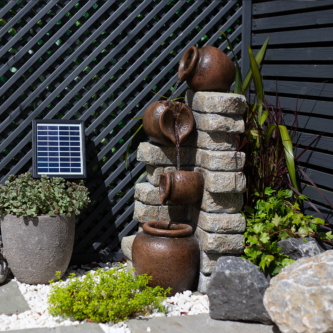 The Cotswold Pours including LEDs by La Hacienda serves as a rustic garden feature, with water cascading through stacked brown pots against a stone wall. This charming setup is solar-powered and beautifully surrounded by lush plants and stones, while a dark lattice fence completes the enchanting backdrop.