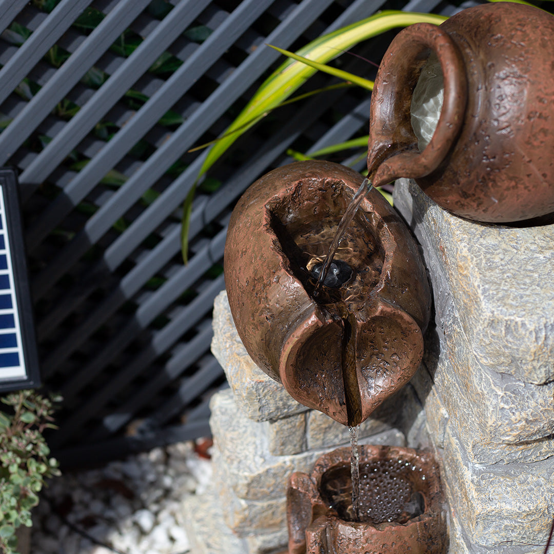 In the Cotswold Pours incl LEDs by La Hacienda, a ceramic teapot pours water onto a cracked, spherical pot in this solar waterfall fountain. With water cascading over stones against a lattice backdrop, it creates the ideal rustic garden feature. A small patch of greenery and part of a solar panel are visible, ensuring energy efficiency throughout the day.