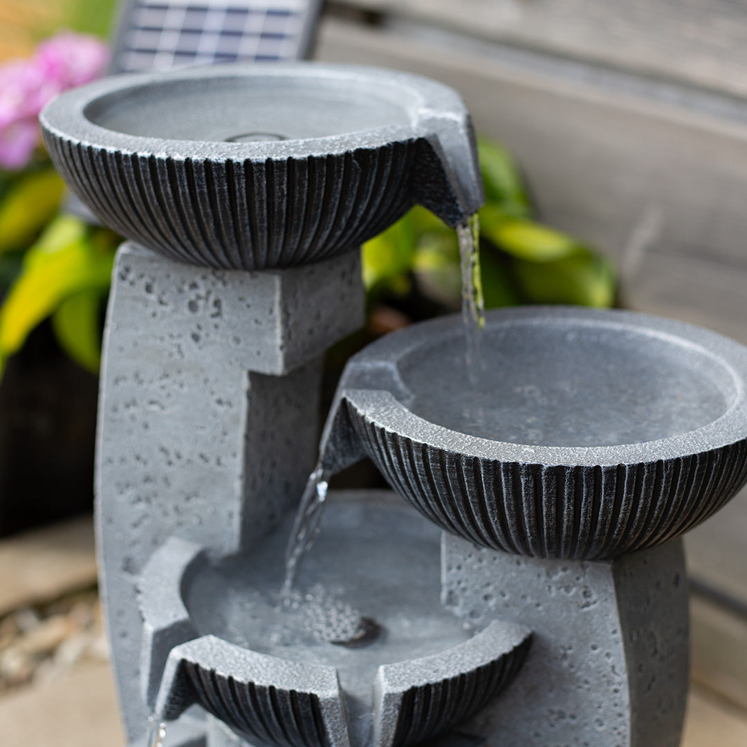 The Greek Columns incl LEDs by La Hacienda serves as a stunning centerpiece in the garden. Featuring a unique three-tiered design, it channels water from one basin to the next, crafting an enchanting cascading effect. Set against a backdrop of lush green plants and a wooden wall, this solar-powered water fountain made of gray stone elevates the elegance of any outdoor space.