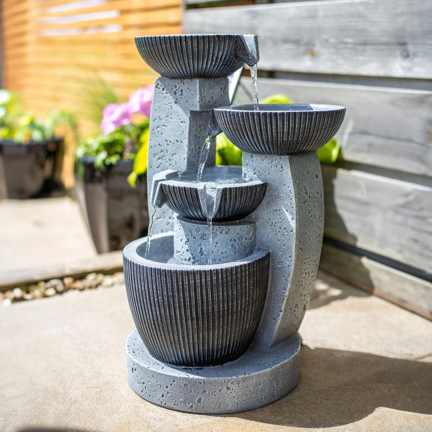 The "Greek Columns incl LEDs" by La Hacienda is a modern solar-powered water fountain featuring three tiered stone bowls that gracefully cascade water from top to bottom. Positioned on a patio with wooden fencing and potted plants, this elegant garden feature adds a serene touch to the outdoor space.