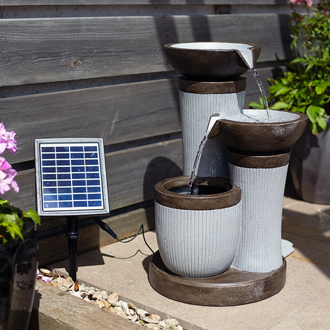 The La Hacienda "Trio Pillars incl LEDs" is a tiered ceramic fountain featuring three bowls and a solar panel on the left. Positioned on a stone patio, this hybrid water feature is encircled by wooden fencing and blooming plants, with water softly cascading from the top bowl to the bottom.