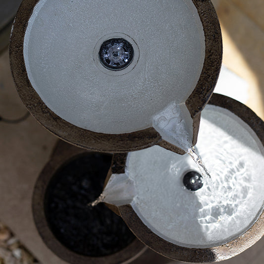 A top-down view of the La Hacienda Trio Pillars incl LEDs reveals a solar-powered waterfall with three tiered stone bowls. Water cascades gently from the top to the bottom bowl, creating a soothing effect as sunlight highlights the texture of the stone and casts shadows along its edges.