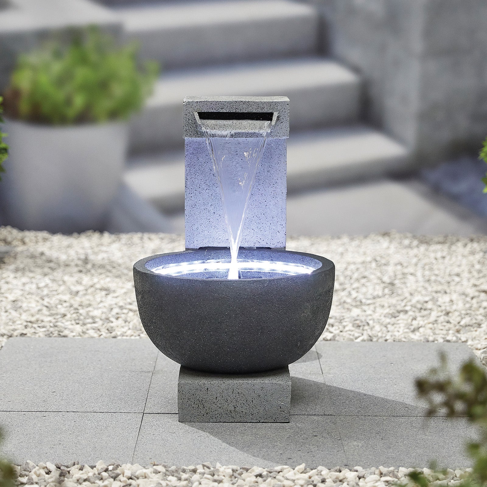 The La Hacienda Solitary Pour (Inc LEDS) Water Feature by La Hacienda is a stunning garden feature, showcasing a bowl-shaped basin with water elegantly cascading from a rectangular spout above. The structure is beautifully illuminated by soft LED lights and is set on a stone tile base surrounded by white gravel, with picturesque plant and stone steps in the background.