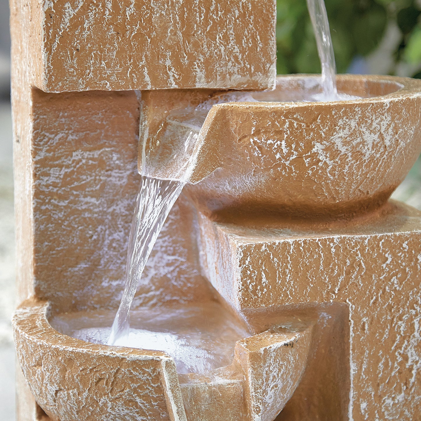 Close-up of the La Hacienda Sparkling Bowls (Inc LEDS) Water Feature, crafted from lightweight poly-resin in textured brown stone. Water cascades from the top tier to the middle and then to the bottom tier, creating a gentle flow. LED lights enhance its charm amidst blurred greenery in the background.