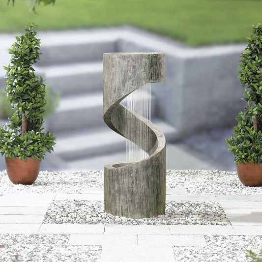 A modern, spiral-shaped outdoor fountain from La Hacienda stands on a gravel surface, bordered by square pavers. The La Hacienda Spiral Showers (Inc LEDS) Water Feature cascades elegantly down thin strands, illuminated by an integrated LED light. Two potted plants with round, green foliage symmetrically frame this self-contained feature.