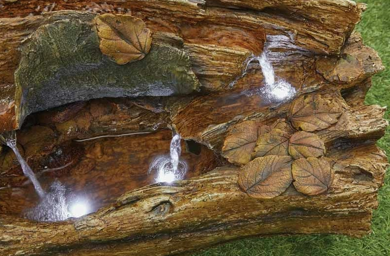 The Balsam Pools inc LEDs by La Hacienda is a decorative water feature designed to resemble a log with multiple cascading streams flowing into a hollow section. It is adorned with artificial leaves and enhanced by LED lights, creating a tranquil ambiance against a grassy background.