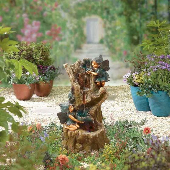 A whimsical garden scene features a fairy sculpture as the centerpiece. The sculpture, nestled within a tree trunk, depicts two fairies surrounded by lush potted plants and colorful flowers. Alongside the distinctive design is the La Hacienda Water Fountain - Distinctive by La Hacienda, with a pathway leading to a garden gate in the distance.