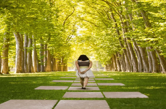 A person walks down a path lined with tall, leafy trees, casting dappled sunlight on the grass. The path, made of Brisks Maestrat Tierra Porcelain Paving with its slip-resistant surface, enhances the serene and peaceful atmosphere.