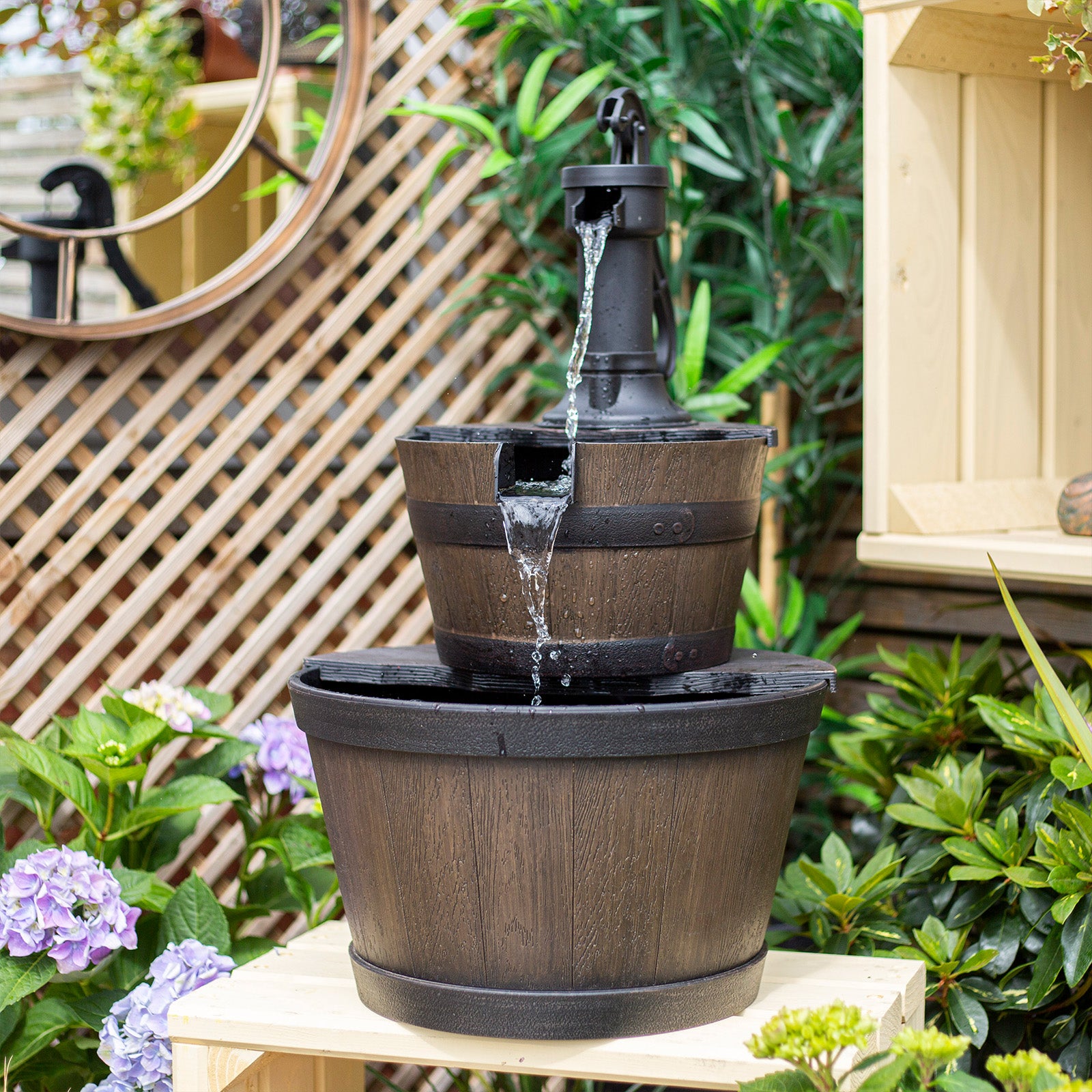 The La Hacienda Whiskey Bowls Water Feature, a tiered wooden barrel fountain by La Hacienda, cascades water from the top spout into two barrels below. This self-contained outdoor water feature is set amid lush greenery and purple hydrangeas in the garden. A lattice wooden backdrop and mirror enhance the aesthetic area.