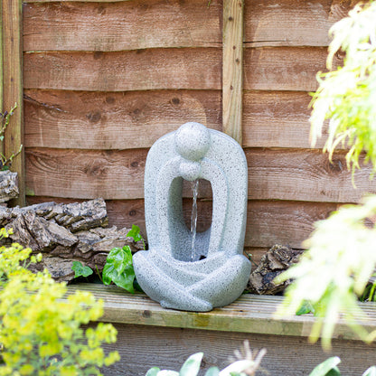 An elegant, tranquil garden water feature with a contemporary, abstract design. Water gently cascades from a sphere into an open figure's lap, framed by a wooden fence and lush green foliage. The La Hacienda Zen Pour Water Feature by La Hacienda is crafted from weather-resistant polyresin and integrates beautifully with the garden ambiance.