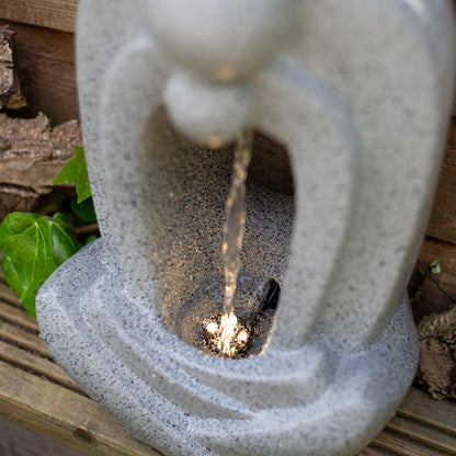 A close-up of the La Hacienda Zen Pour Water Feature by La Hacienda reveals a continuous stream of water flowing from the top, illuminated by LED lights at the base. The weather-resistant polyresin fountain boasts a modern, abstract design and is placed outdoors near a wooden fence with some green leaves partially visible.