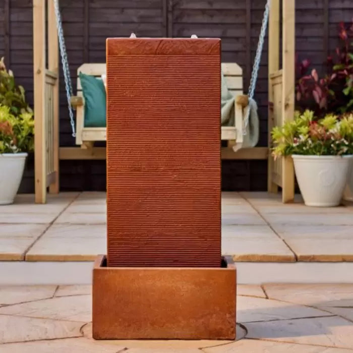 The Amber Falls by La Hacienda, a contemporary oxidized vertical rectangular terracotta garden water feature, graces the patio. Behind it, a wooden swinging chair with cushions and potted plants create a serene setting. This self-contained structure stands out against the backdrop of a dark wooden fence.