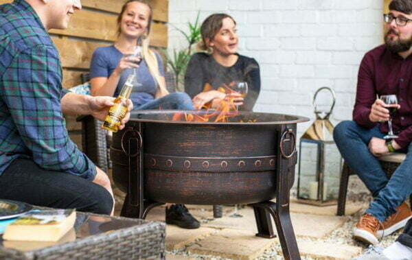 Four people gather around the La Hacienda Brava Firepit, featuring a stylish bronze effect, as they relish drinks and engage in conversation. The ambiance is cozy, with a brick wall, potted plant, and lanterns in the background enhancing their relaxed and social atmosphere.