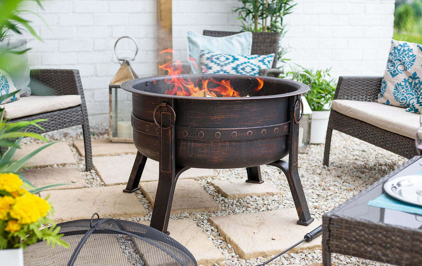 A cozy outdoor patio features the La Hacienda Brava Firepit, showcasing flames dancing in a bronze effect. It's surrounded by wicker seating adorned with blue and white patterned cushions. Decorative plants and a lantern accent the space, while yellow flowers add a vibrant pop of color.