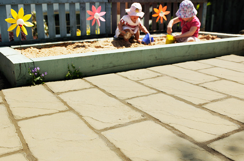 Two young children are playing in a sandbox situated in a fenced outdoor area. They are wearing sun hats and sitting on the sand, engaging with toys. Colorful pinwheel decorations are placed around the sandbox. The ground is paved with Brisks' Weathered York Cathedral Paving Kit by Bowland Stone, adding charm to the setting.