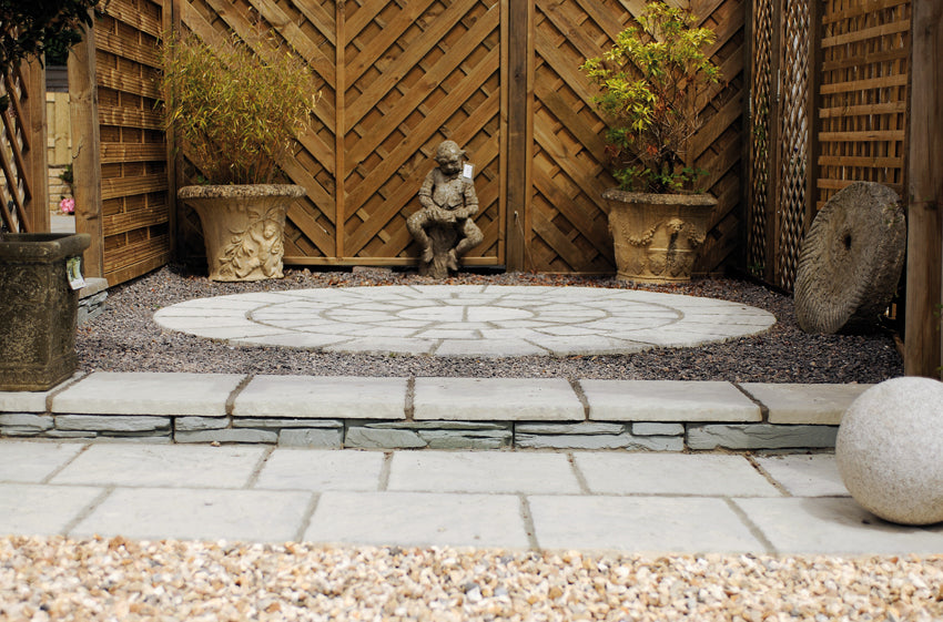 A serene garden features a circular stone patio with a contemplative statue of a seated person at the center. The patio, crafted from the Weathered Moss Cathedral Paving Kit by Bowland Stone, is surrounded by wooden lattice panels and potted plants, with gravel and a large round stone at the edges. The paving kit is provided by Brisks.