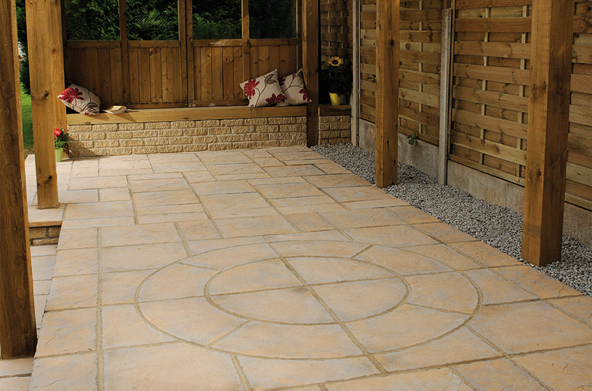 A patio area featuring Welsh Slate Chalice Circle tiles by Bowland Stone, arranged in a geometric pattern. Wooden fencing, rich in honey brown hues, lines the right side, while a wooden bench with pillows is positioned at the back. Potted plants and a sunflower on a stand are next to the bench. The area is shaded by a wooden pergola.