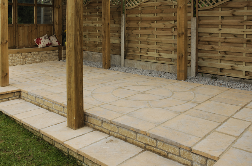 A backyard patio featuring light beige stone tiles, with a Welsh Slate Chalice Circle by Bowland Stone from Brisks etched in the center. Wooden posts support an overhead structure, and a fence with lattice tops borders the area. In the corner, there is a cushioned wooden bench with floral pillows and charming garden features.