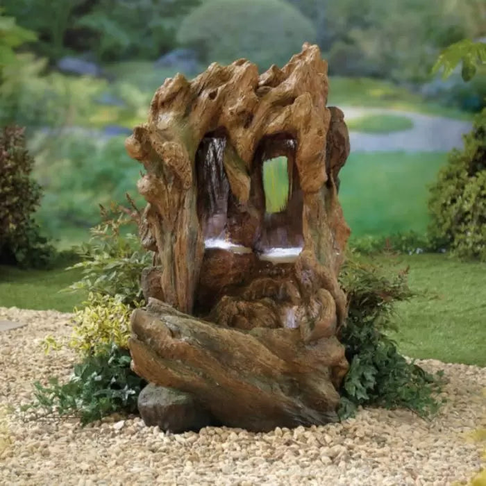 The Colorado Falls inc LEDS by La Hacienda is a stunning garden water feature designed to emulate a tree trunk, complete with two cascading water streams. Surrounded by lush greenery and pebbles, it seamlessly enhances any garden environment.