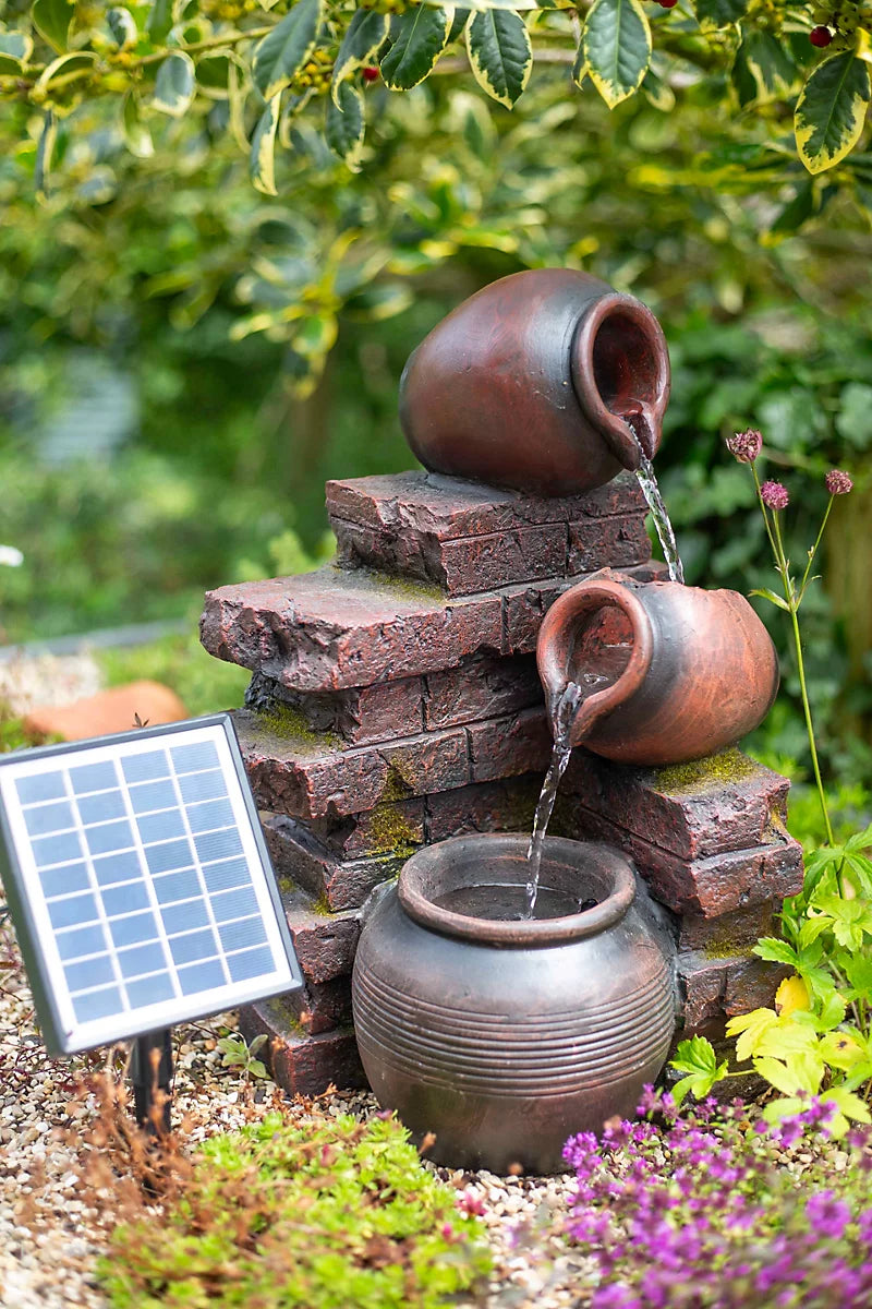 In a garden scene, the La Hacienda Pot Wall Pours solar-powered water feature showcases a tiered fountain with three clay pots gently pouring water over stacked bricks into a large pot. Surrounded by lush greenery and purple flowers, it embodies tranquility and enhances the outdoor space's eco-friendly charm.