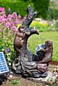 The decorative solar water fountain, branded as La Hacienda's "Woodland Otters," showcases two playful otters climbing a tree. Water cascades from a spout into a rock basin, surrounded by vibrant pink flowers and lush green foliage, creating an enchanting woodland setting.