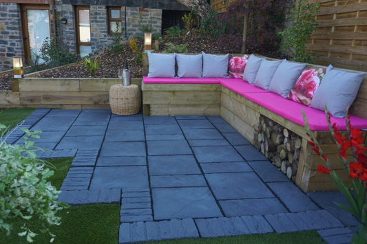 A cozy outdoor seating area features a wooden L-shaped bench with pink cushions and floral pillows, positioned against a wooden fence. The space is paved with the Welsh Slate Chalice Paving Kit by Bowland Stone from Brisks, surrounded by greenery and illuminated by modern outdoor lights, making it one of the standout garden features.