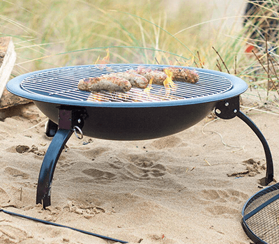 A La Hacienda Firepit - Explorer featuring a metal grate is set up on a sandy beach, with grass visible in the background. The portable firepit from La Hacienda is grilling three sausages over an open flame.