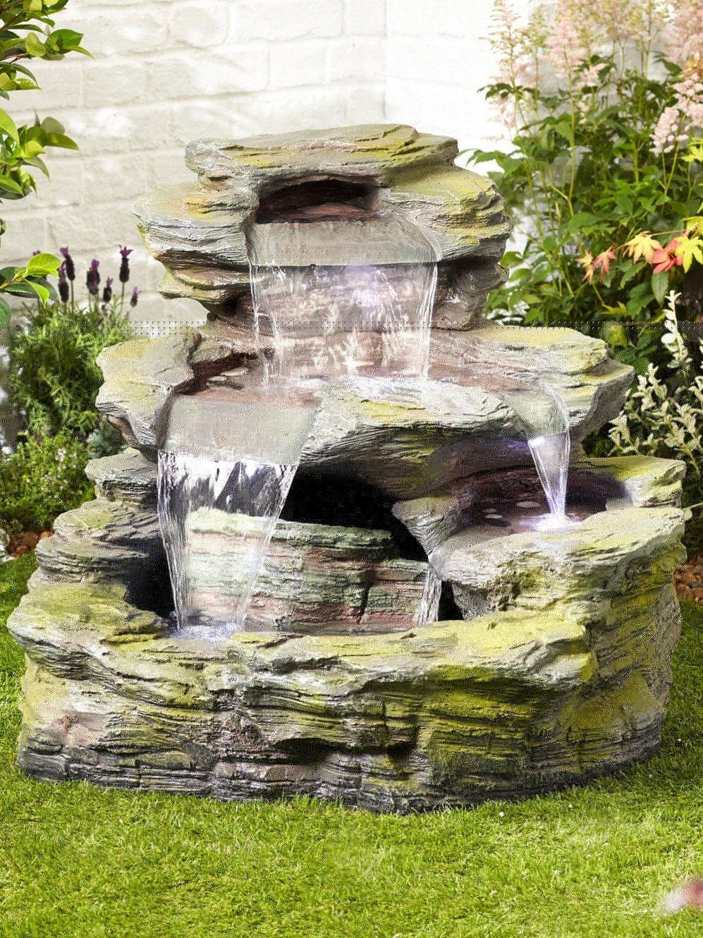 A decorative three-tiered water feature from La Hacienda, the Garda Falls inc LEDS is crafted from durable resin and features waterfalls cascading down each level. Nestled in a lush garden with green grass and various plants, it is beautifully illuminated by LED lights to enhance its charm even at night.