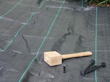 A wooden mallet and black plastic pegs sit on a GROUNDTEX Woven Geotextile Membrane 1x15 from Growtivation, a black weed barrier fabric with green lines, laid and partially unfolded near plants.