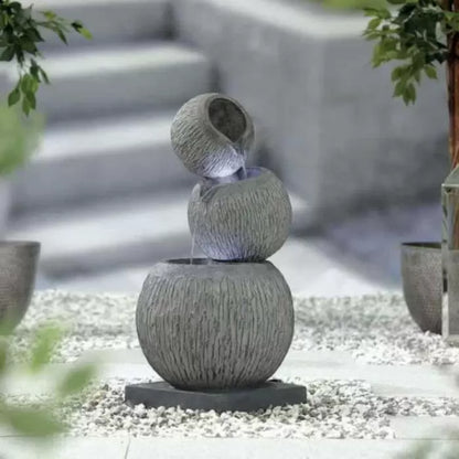 The Illusion Falls inc LEDs by La Hacienda is an ornamental outdoor fountain featuring three stacked, textured stone bowls. Water gracefully flows from the top bowl to the bottom, complemented by soft LED lighting. This poly-resin design is encircled by white pebbles and accompanied by potted plants.