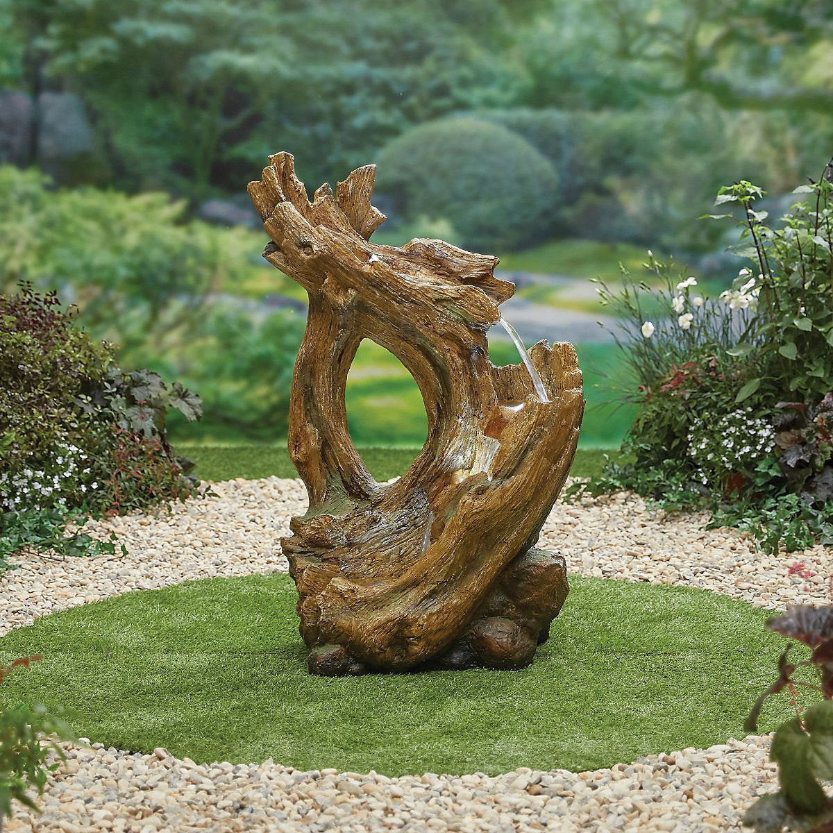 The Knotted Willow Falls sculpture by La Hacienda stands on a small grassy patch, surrounded by a ring of pebbles. The serene garden setting, enhanced by lush greenery and trees, now includes LED lights that subtly illuminate the sculpture at night.