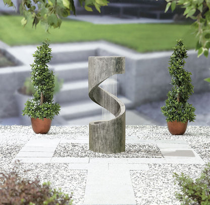 The La Hacienda Spiral Showers inc LED lights elegantly twists on a gravel surface, flanked by two potted topiary trees. As daylight fades, the integrated LED lights softly illuminate the scene, while steps and greenery whisper in the blurred background.