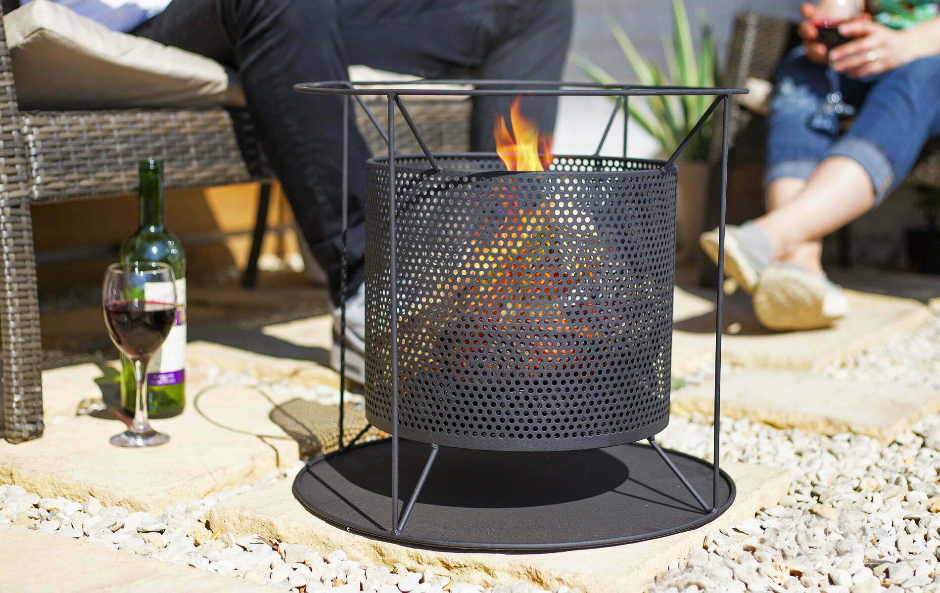 The La Hacienda Kora Fireplace, a round metal piece with a perforated design, is lit and placed on a stone patio. Nearby, two people unwind on wicker furniture with a bottle of wine and glasses beside them, creating a cozy outdoor ambiance accentuated by its contemporary design.