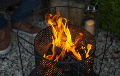 A small outdoor fire pit, designed with contemporary flair, presents bright orange flames dancing on burning logs atop white gravel. A candle flickers in a lantern nearby as a person's legs in jeans and brown shoes rest beside the scene, savoring the warmth provided by La Hacienda's Kora Fireplace's durable steel construction.