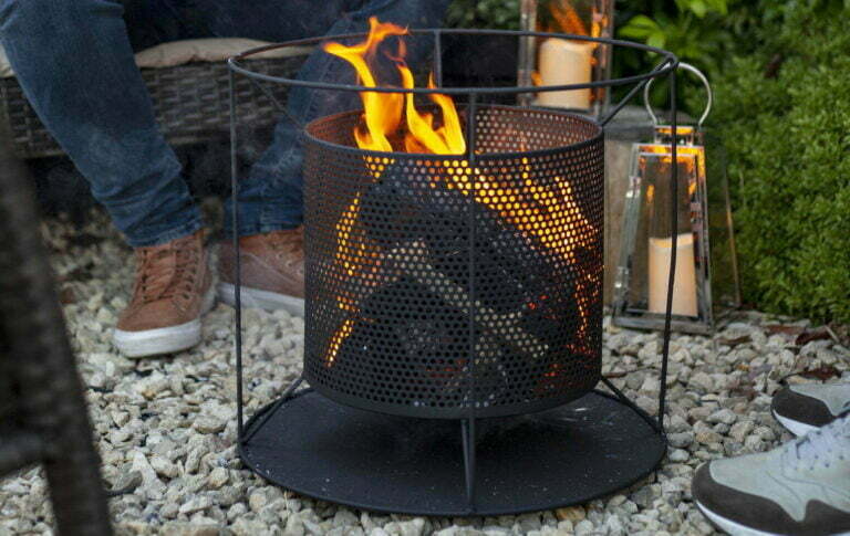 A cozy fire pit with burning logs inside the La Hacienda Kora Fireplace, crafted from durable steel construction. It rests within a metal mesh container, surrounded by gravel. Two people are seated nearby, sneakers visible. Lit lanterns enhance the warm ambiance with a touch of contemporary design.