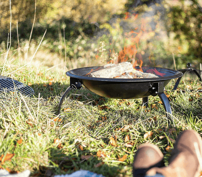 A La Hacienda Firepit - Explorer with burning logs sits on a grassy outdoor area. The fire emits vibrant flames, surrounded by scattered autumn leaves. In the foreground, a person’s crossed legs are visible, suggesting they are relaxing while enjoying the La Hacienda Firepit - Explorer.