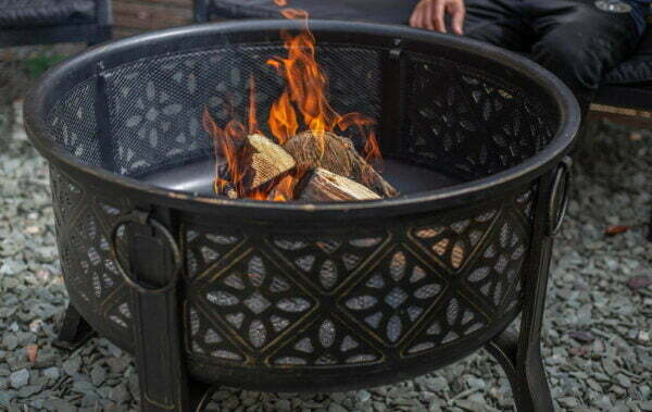 A La Hacienda Moresque Firepit from the brand La Hacienda, showcasing detailed Moresque designs, is ablaze with wood. The lively flames emit a bright, warm glow. Positioned on a gray gravel bed, the ambiance radiates Moroccan style with a subtle glimpse of a person’s leg in the background.