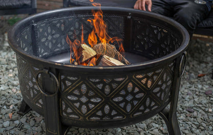 The La Hacienda Moresque Firepit is a decorative outdoor fire pit designed to burn wood, showcasing circular metal mesh patterns. The flames that rise above the logs give off North African vibes. Grey stones cover the ground, and part of a person's leg can be seen in the background.