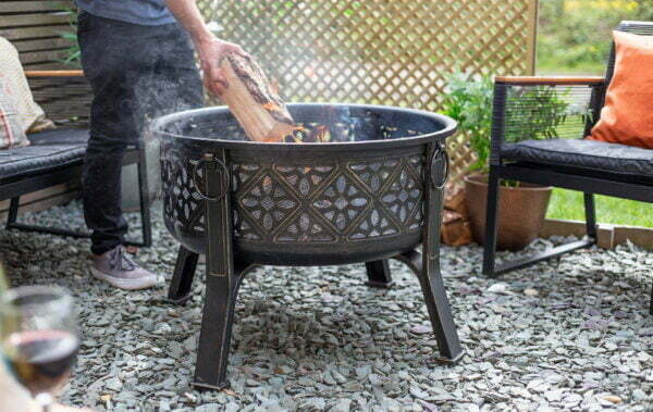 Using the La Hacienda Moresque Firepit, a person adds a log to the outdoor firepit, which exudes North African vibes. The surrounding area is paved with stones and features chairs and a wooden privacy screen. In the foreground, an elegant glass of red wine completes the scene.