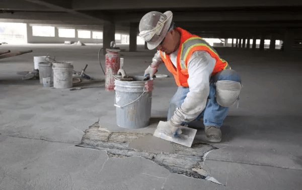 A construction worker in a high-visibility orange vest, safety helmet, and knee pads is repairing a cracked concrete floor. The worker is using a trowel to apply pre-blended Instant Concrete mix from a bucket. Several buckets of Brisks Instant Concrete and other tools are visible in the background.