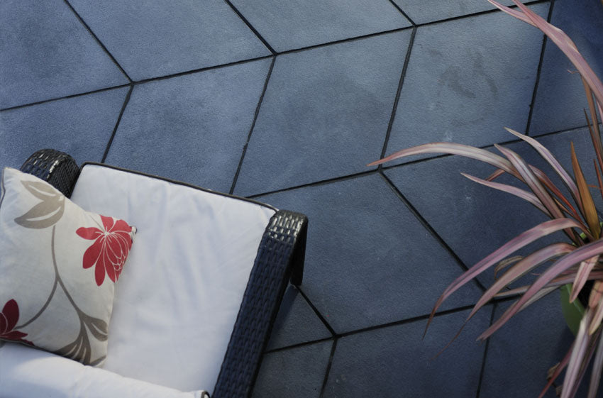 A top-down view of a patio showcases dark, geometric pavers from the Welsh Slate Prestbury Chevron Patio Kit by Bowland Stone under the Brisks brand. In the lower left corner, a wicker chair with a white cushion and a pillow featuring red and grey floral patterns is placed. On the right side, part of a plant with long, slender leaves can be seen.