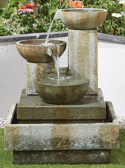 The Patina Bowls by La Hacienda is a tiered outdoor water fountain made from glass fiber reinforced concrete. It includes three stone basins with cascading water, designed to create a tranquil oasis amidst a garden lush with flowers and greenery.