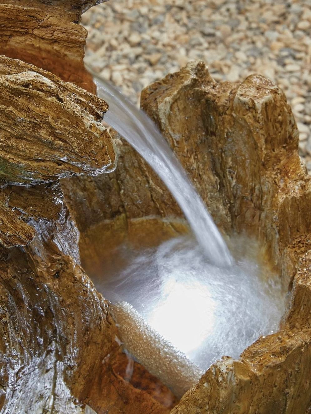 The Knotted Willow Falls with LEDs by La Hacienda features a close-up view of water cascading over textured, rocky surfaces into a small pool. Enhanced by LED lights that illuminate the scene, this piece showcases natural stone formations with a layered appearance. The water creates a clear, reflective surface in the pool while pebbles are visible in the background.